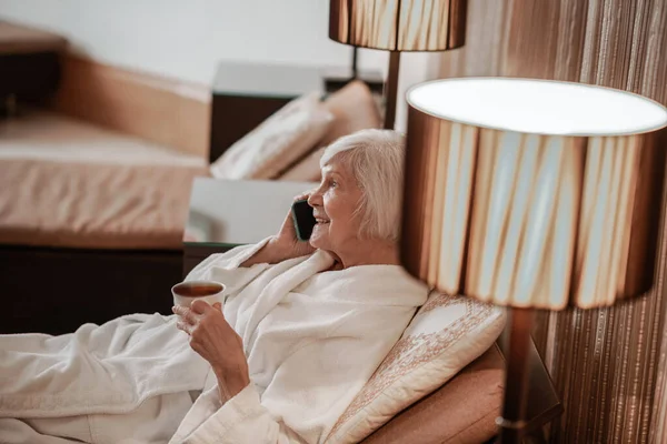 Uma mulher de cabelos grisalhos sênior deitada na cama e tomando café da manhã — Fotografia de Stock