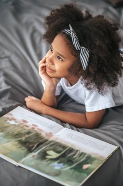 A cute girl with hair band lying on bed with a book