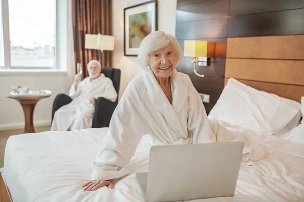 An old woman with a laptop sitting on a bed in a hotel room — Foto Stock