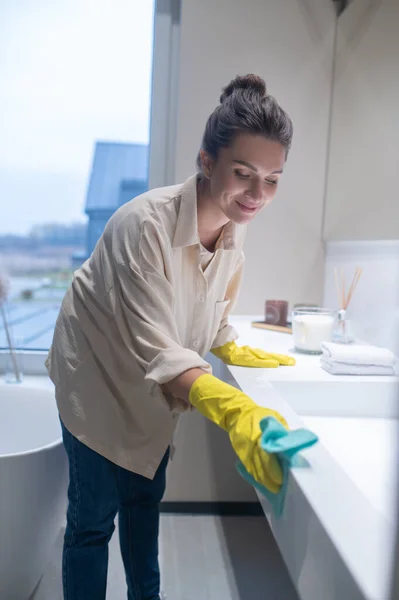 Молода жінка в жовтих рукавичках прибирає поверхні у ванній — стокове фото
