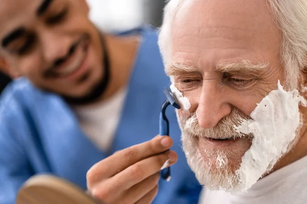 Gioioso caregiver rasatura un uomo anziano con un rasoio — Foto Stock