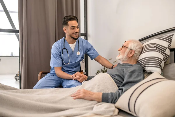 Веселий медичний працівник втішає старого, який лежить у ліжку — стокове фото