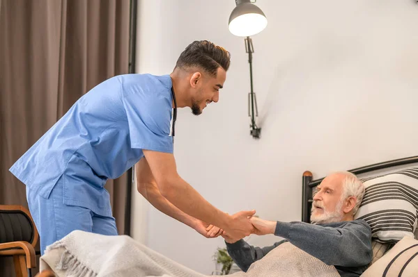 Усміхнений медичний працівник піднімає старого з ліжка — стокове фото