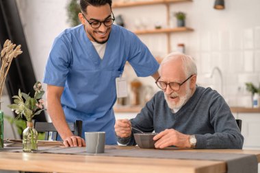 Joyous caregiver watching the pensioner eating breakfast