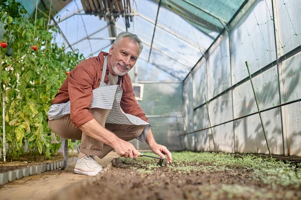Kameraya bakan adam sera bahçesinin yanında çömelmiş.