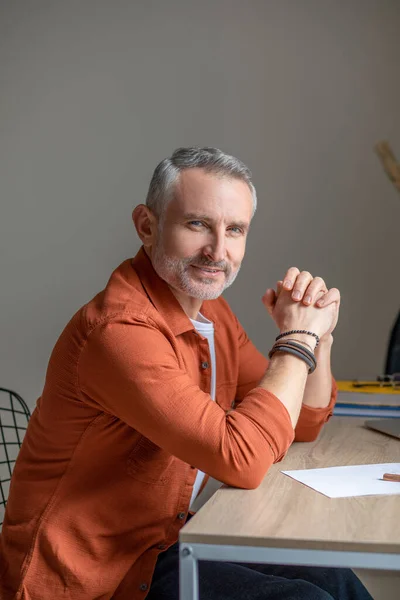Een man in oranje shirt zit aan tafel en kijkt tevreden — Stockfoto