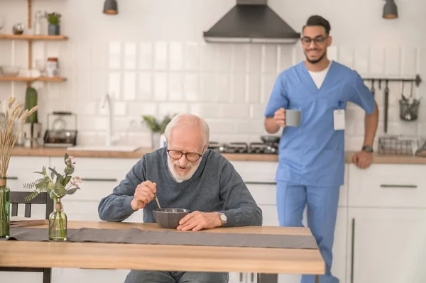 Viejo comiendo en presencia de su alegre cuidador — Foto de Stock