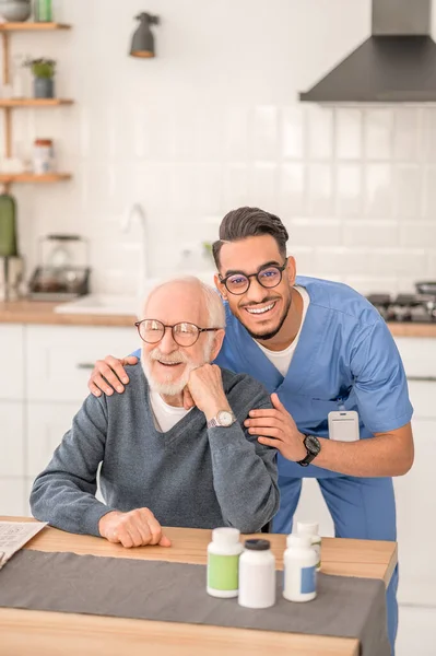 Cuidador alegre abrazando a un pensionista sentado en la mesa de la cocina — Foto de Stock