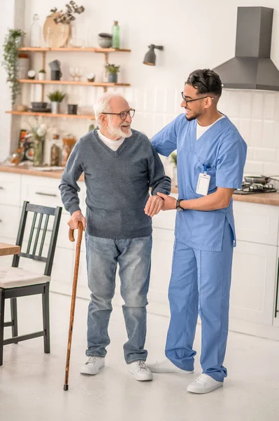 Joyous pensioner moving around the kitchen assisted by his caregiver — Stock Photo, Image