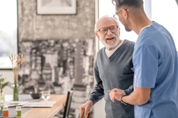 Old man standing by the table supported by a nurse — Stock Photo, Image