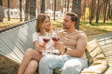 Gülümseyen romantik genç erkek hamakta yanında oturan güzel kız arkadaşına kadeh kaldırıyor.