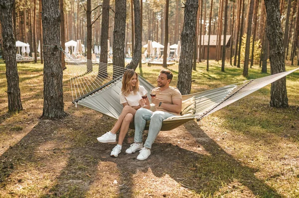 Smiling young woman seated in the hammock beside her joyful boyfriend toasting to their relationship
