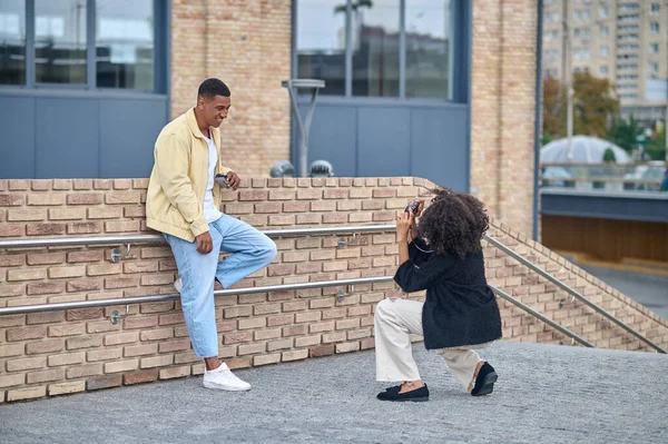 Yürüyorum, fotoğraf çekiyorum. Kıvırcık saçlı kız çömelmiş fotoğraf çekiyor koyu tenli adam poz veriyor bacağı şehir caddesinde eğilmiş duruyor