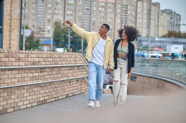 Good view. Enthusiastic dark-skinned guy gesturing with hand to side and looking surprised girl with camera on neck walking in city