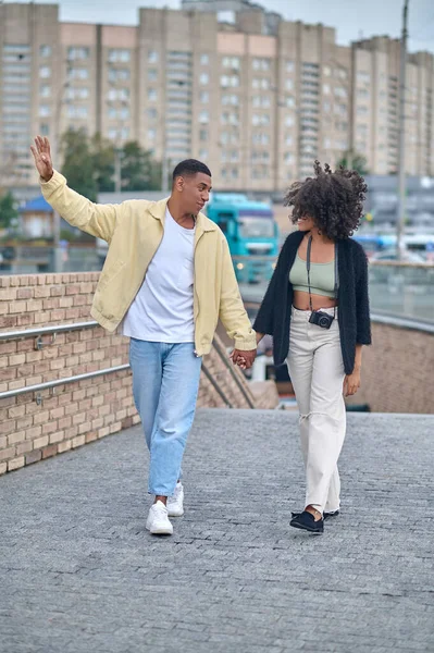 Happy people. Happy dark-skinned guy gesturing and girl with camera walking hand in hand looking at each other on street
