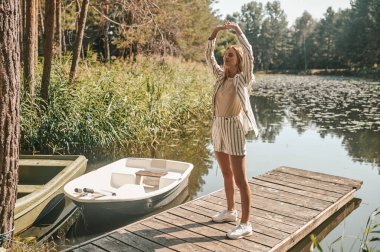 Ahşap bir iskelede, gözleri kapalı ve kolları havada duran sakin bir kadının tam boy portresi.