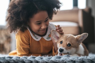 Arkadaşız. Turuncu elbiseli kıvırcık saçlı bir kız yavru köpekle oynuyor.