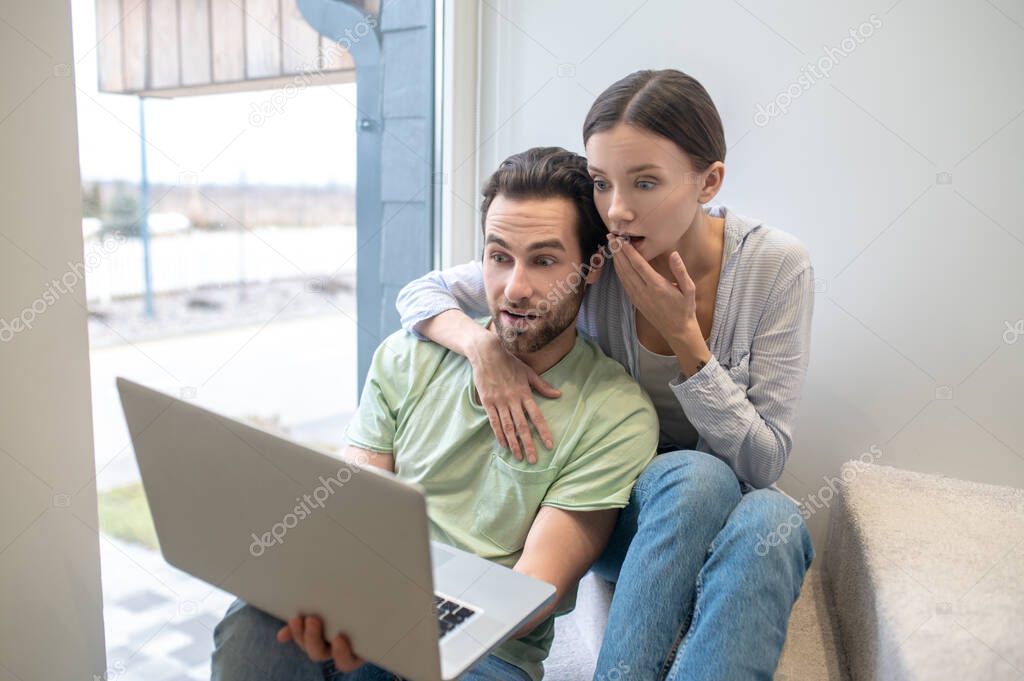 Surprise, question. Young adult man and woman touching hand to mouth questioningly surprised looking at laptop sitting near window indoors