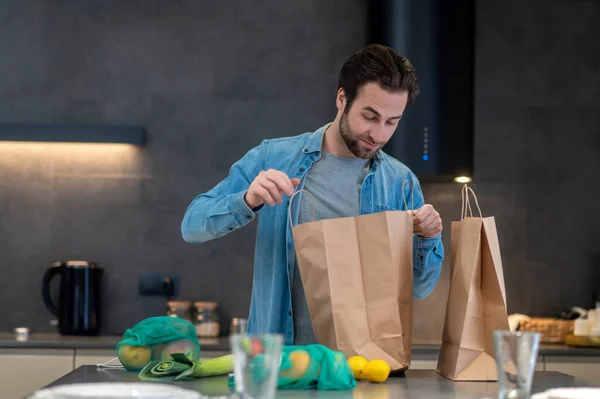 Interest. Bearded young man interested looking into paper bag with groceries standing on table in modern kitchen at home