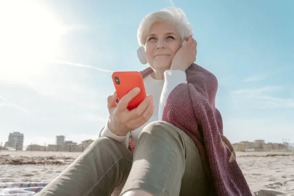 ワイヤレスヘッドフォンと砂の上に座って手にスマートフォンで明るい高齢者の女性を笑顔 — ストック写真
