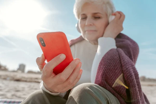 砂の上に座ってスマートフォンの画面を見ている無線ヘッドフォンのシニア女性 — ストック写真