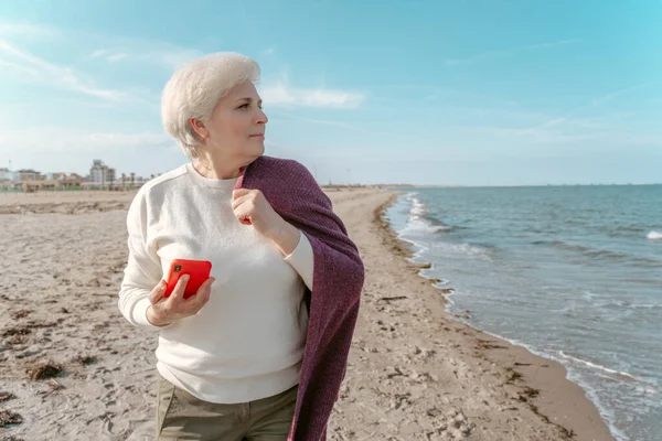 ビーチに立って離れて見て手にスマートフォンを持つ思慮深い美しい高齢者の女性 — ストック写真