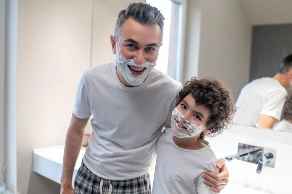 Funny shaving. Dad and son looking funny with shaving foam on their faces