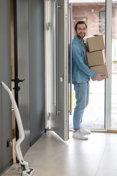 Delight. Young bearded man with boxes in his hands entering new house looking enthusiastically into room