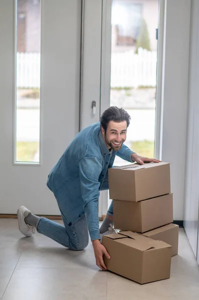 Lucky day. Smiling bearded man looking at camera crouching near boxes gathering them together in hallway of new apartment