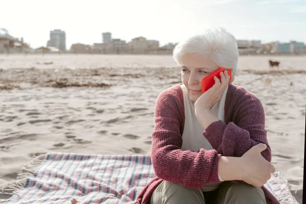 砂浜に一人で座って スマートフォンを呼び出す穏やかな魅力的なシニア女性 — ストック写真