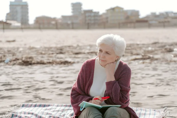 ボールポイントのペンと砂の上に座って開いた日記を持つ思慮深い白髪の女性が離れて見て — ストック写真