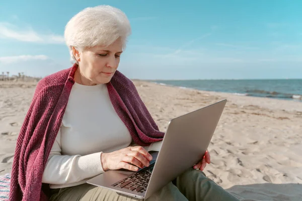 深刻な集中魅力的な白人の白髪の女性はビーチに座って ラップトップを入力します — ストック写真