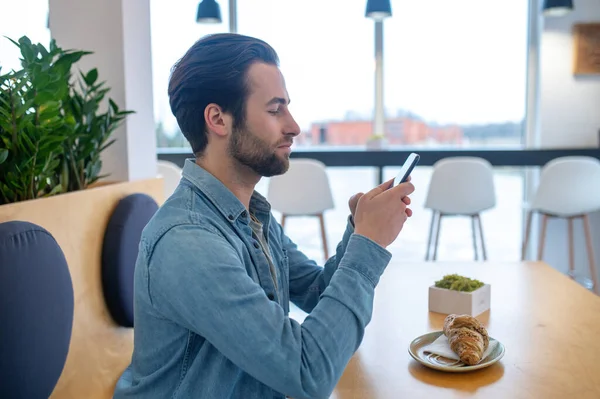 Important information. Concentrated man sitting at table sideways to camera looking interestedly into smartphone in cafe during day