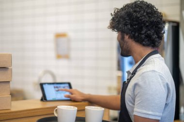 Cafe, sipariş ver. Önlüklü kıvırcık saçlı bir adamın tablet ekranına dokunduğu tabletin yanındaki tepside ayakta duruşu.