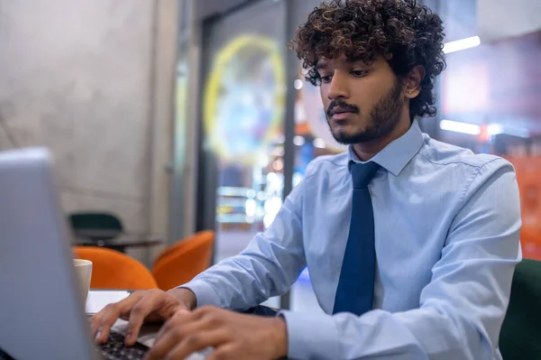 Typing, job. Young indian mustachioed man in tie concentrated typing on laptop sitting at table indoors