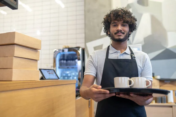 Waiter. Happy indian curly haired man in apron with tray walking towards camera near counter in cafe