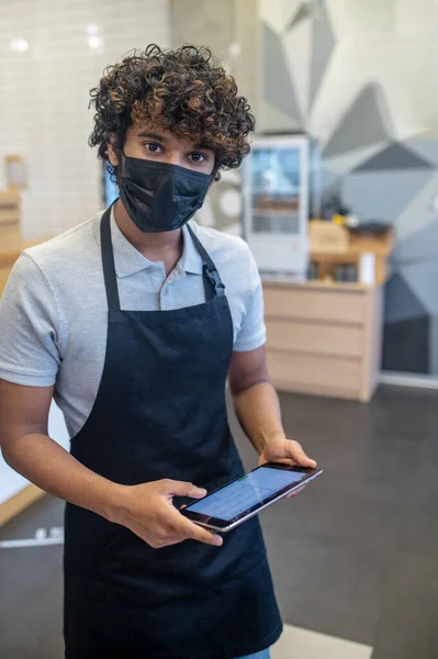 Good service. Indian friendly curly haired man in protective mask and apron holding tablet looking at camera standing in cafe