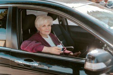 Motorlu araçta oturan gri saçlı kadın tablet bilgisayar ve tek kullanımlık kahve bardağıyla.