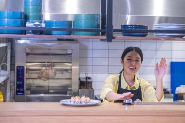 Sipariş hazır. Önlüklü Asyalı kadın dikkat çekiyor kafeteryada suşi tabağının yanında çana dokunuyor.