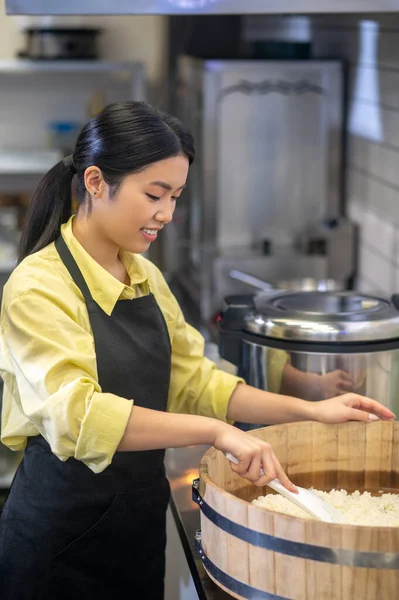 漂亮的穿着围裙的亚洲姑娘站在大碗旁 用勺子在咖啡厅里准备食物 — 图库照片