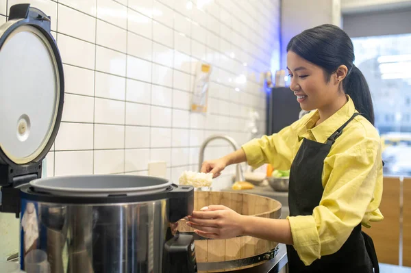 Work, pleasure. Cheerful asian woman in apron holding rice spoon over bowl standing near stove at workplace in cafe