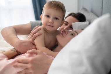 Şefkat. Genç bir kadın ve bir adam yatakta uzanmış kameraya bakarken küçük şirin bir bebeğe sarılıyorlar.