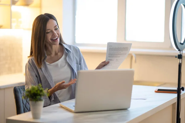 Self-employment. Cute young woman pointing at papers smiling at laptop while sitting at table at home during daytime