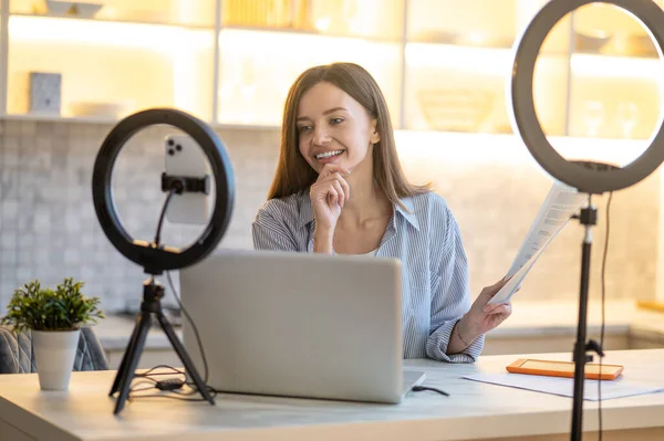 Discussion. Young smiling woman with papers touching chin thinking looking at laptop sitting at table at home