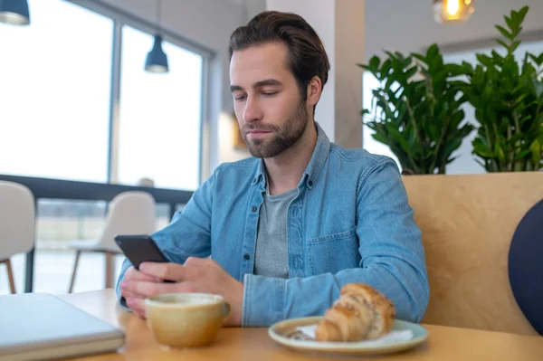 Interesting here. Young bearded man interested reading message looking at smartphone screen sitting at table with cappuccino and croissant