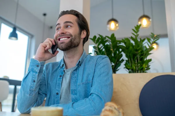 Phone call. Smiling young bearded man looking away talking on smartphone sitting indoors during day