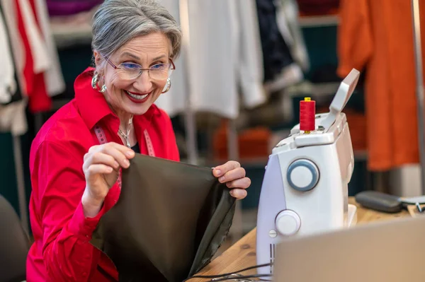 Fabric for sewing. Excited smiling woman in glasses sitting at sewing machine showing fabric at laptop screen in atelier