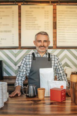 Küçük bir iş. Gri saçlı, önlüklü, sakallı, kendine güvenen bir adam, kafenin tezgahında kameraya bakıyor.