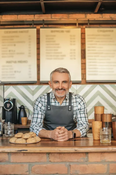 Welcome. Friendly middle aged man in apron standing behind bar with pastries smiling at camera in his cafe
