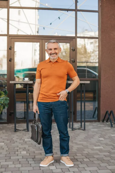 Lucky day. Gray-haired attractive man with suitcase standing outdoors against backdrop of city building smiling at camera